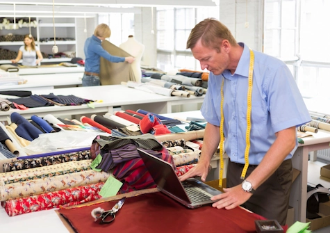 Empresa textil con muchas telas, un hombre con un ordenador y un metro colgando, que representa el sastre y de fondo mujeres con telas trabajando